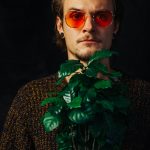 man in black and white long sleeve shirt wearing sunglasses standing beside green plant